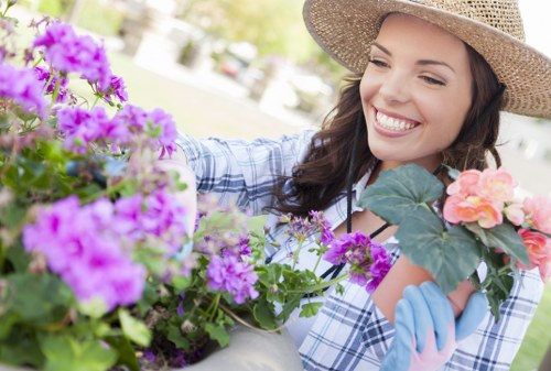 A woman in a wide-brimmed straw hat and light blue gloves is tending to a vibrant flower arrangement in a garden. She is smiling as she looks at the colorful blooms, which include pink, purple, and peach flowers, with lush green leaves surrounding them. The garden setting features a well-maintained lawn in the background, along with a variety of flowering plants and shrubs, all bathed in natural sunlight. The scene suggests activities such as planting or arranging flowers, reflecting professional gardening and outdoor maintenance services provided by Lawn Mowing Staines in the Staines area, with a focus on creating attractive and well-kept outdoor spaces. The environment appears peaceful and flourishing, highlighting the importance of garden care in enhancing outdoor aesthetics in the local area.