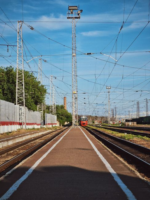 A series of railway tracks running parallel across a yard, with gravel and soil between them, leading towards industrial freight cars lined up on distant sidings. Overhead, there is a network of red a